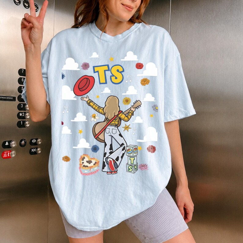 May include: A light blue t-shirt featuring a graphic of a person with blonde hair wearing a yellow plaid shirt and cow-print trousers, holding a guitar and a red hat. The design includes white clouds, flowers, blocks with the letters TS, and the text TS.