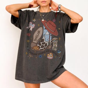 May include: A charcoal gray short-sleeve t-shirt featuring a graphic print of a cow-print cowboy boot with the name Taylor, a brown cowboy hat, stacked blocks with the letters T, S, and 13, a bow, a heart, and a beaded chain with stars.