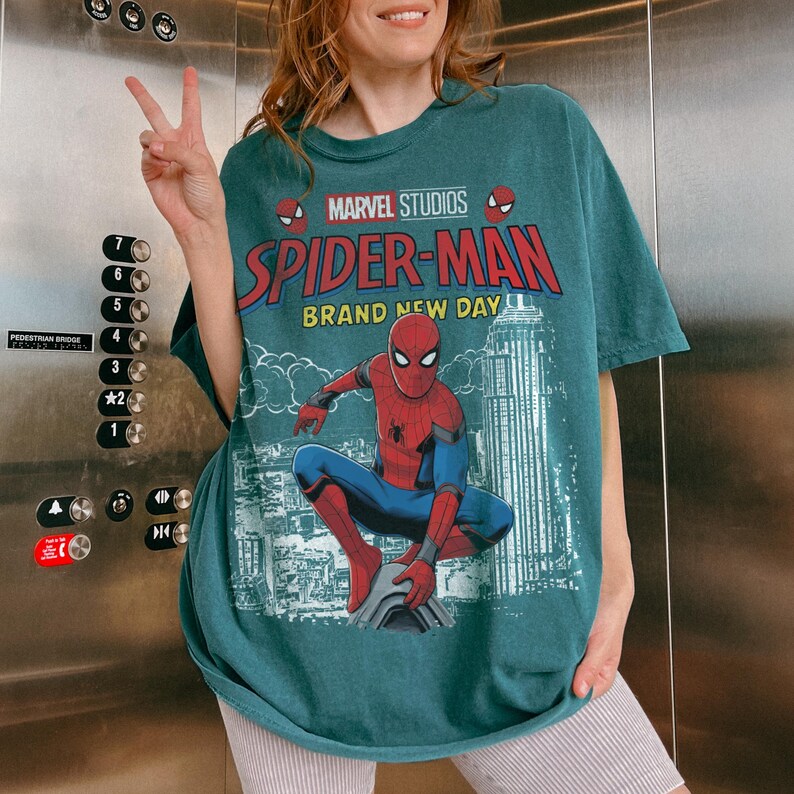 May include: A teal short-sleeve t-shirt featuring a graphic of Spider-Man in a crouching pose. The text reads Marvel Studios Spider-Man Brand New Day. The design includes a city skyline background in white and red accents on the character suit.