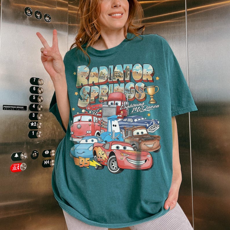 Op de afbeelding: Een donkergroenblauw t-shirt met korte mouwen en een grafische print van Radiator Springs uit de film Cars. Het ontwerp bevat diverse voertuigpersonages, een gouden trofee en de tekst Radiator Springs en Lightning McQueen in een retro lettertype.
