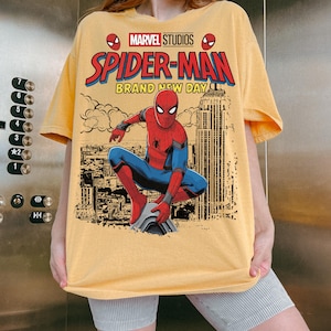 May include: A yellow t-shirt featuring a graphic print of Spider-Man crouching over a city skyline. The text reads Marvel Studios Spider-Man Brand New Day. The design includes a red and blue Spider-Man figure and a black and white city background.