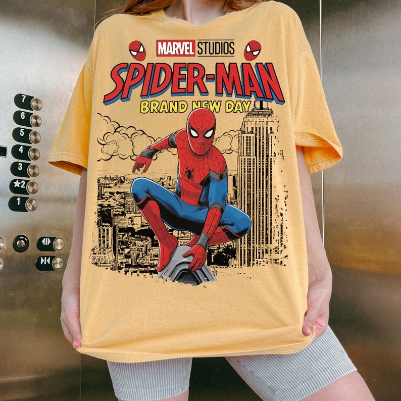 May include: A yellow t-shirt featuring a graphic print of Spider-Man crouching over a city skyline. The text reads Marvel Studios Spider-Man Brand New Day. The design includes a red and blue Spider-Man figure and a black and white city background.