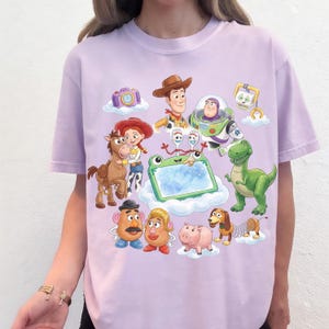 May include: A light purple t-shirt featuring a colorful graphic of characters from the animated movie Toy Story. The design includes Woody, Buzz Lightyear, Jessie, Rex, and other characters on a cloud background.