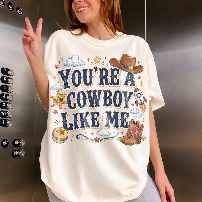 May include: An off-white t-shirt featuring the text "YOU'RE A COWBOY LIKE ME" in dark blue letters. The graphic includes a brown cowboy hat, a yellow sheriff star, a rocket ship, a pair of brown cowboy boots, a colorful ball, and small clouds.