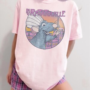 May include: A light pink t-shirt featuring a graphic from the movie "Ratatouille." The graphic shows a blue rat wearing a chef's hat in front of a Parisian scene with the Eiffel Tower. The text "La Ratatouille" is above the graphic.