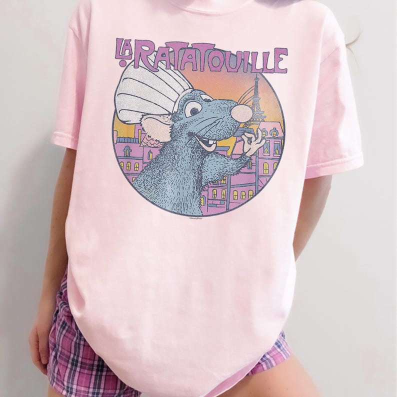 May include: A light pink t-shirt featuring a graphic from the movie "Ratatouille." The graphic shows a blue rat wearing a chef's hat in front of a Parisian scene with the Eiffel Tower. The text "La Ratatouille" is above the graphic.