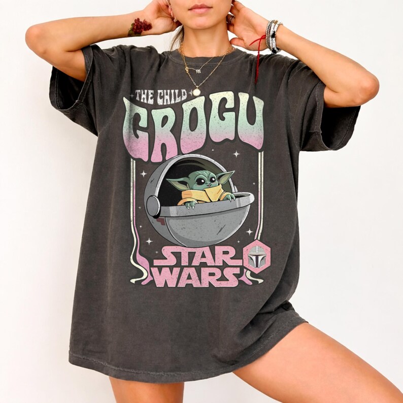 K&ouml;nnte beinhalten: Ein anthrazitfarbenes Kurzarm-T-Shirt mit einer Grafik von Grogu in einer schwebenden Kapsel. Der Text lautet THE CHILD GROGU und STAR WARS in einer Retro-Schriftart mit rosa und gr&uuml;nen Farbverl&auml;ufen. Das Design enth&auml;lt kleine Sterne.
