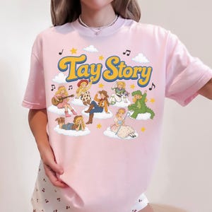 Puede incluir: Una camiseta rosa claro con el texto Tay Story en amarillo con borde azul. El gr&aacute;fico incluye varias ilustraciones de un personaje rubio con diversos atuendos, como una vaquera, un traje espacial y un disfraz de dinosaurio, sobre nubes blancas con estrellas amarillas y notas musicales negras.