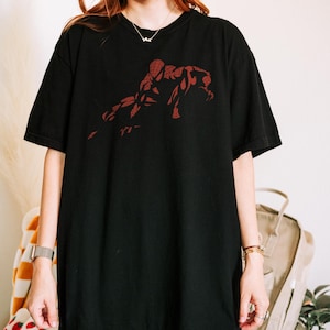 May include: A black short-sleeved t-shirt featuring a dark red graphic print of a crouching superhero figure on the chest. The shirt has a crew neckline and a relaxed fit, showcasing a detailed silhouette of the character in a dynamic pose.