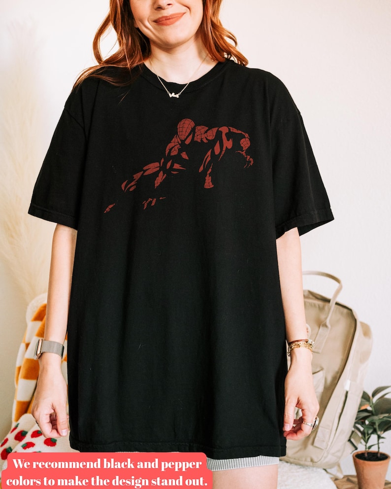 May include: A black short-sleeved t-shirt featuring a dark red graphic print of a crouching superhero figure on the chest. The shirt has a crew neckline and a relaxed fit, showcasing a detailed silhouette of the character in a dynamic pose.