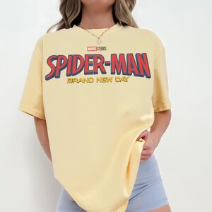 May include: A pale yellow short-sleeve t-shirt featuring the Marvel Studios logo and the text Spider-Man Brand New Day in large red and blue lettering across the chest. The shirt has a crew neckline and a relaxed, loose-fitting silhouette.