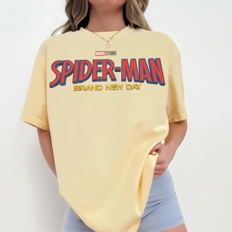 May include: A pale yellow short-sleeve t-shirt featuring the Marvel Studios logo and the text Spider-Man Brand New Day in large red and blue lettering across the chest. The shirt has a crew neckline and a relaxed, loose-fitting silhouette.