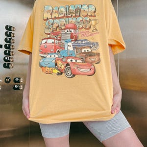 Op de afbeelding: Een geel t-shirt met korte mouwen en een grafische print van personages uit de film Cars. Het ontwerp bevat de tekst Radiator Springs en Lightning McQueen, samen met illustraties van diverse voertuigen en een gouden trofee.