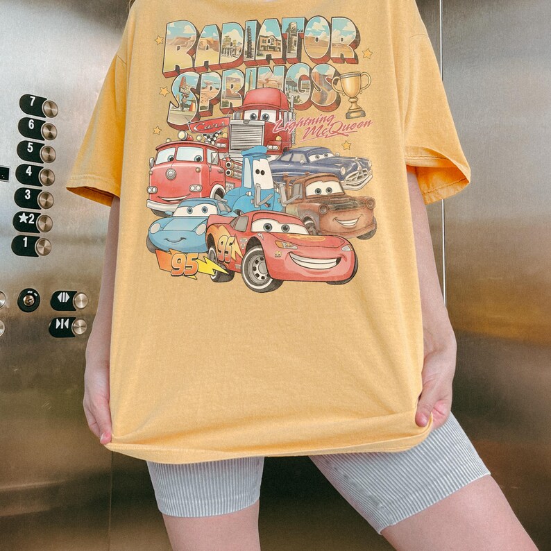 Op de afbeelding: Een geel t-shirt met korte mouwen en een grafische print van personages uit de film Cars. Het ontwerp bevat de tekst Radiator Springs en Lightning McQueen, samen met illustraties van diverse voertuigen en een gouden trofee.