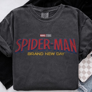 May include: A charcoal gray short-sleeve t-shirt featuring the red and blue text Spider-Man, the yellow text Brand New Day, and the Marvel Studios logo printed on the chest. The shirt has a crew neckline and a faded, vintage-style fabric finish.