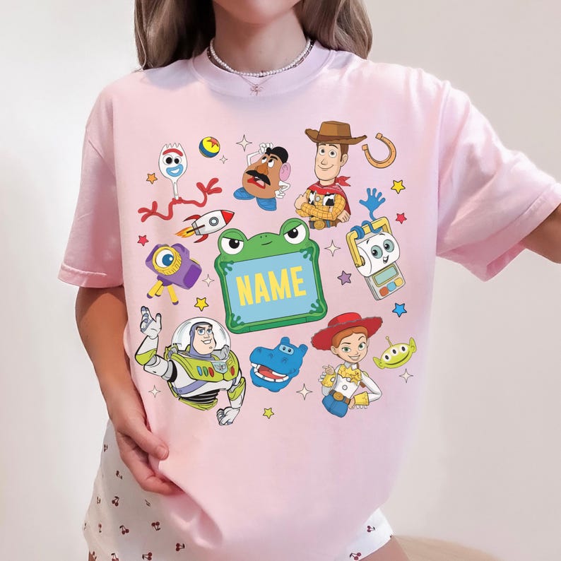 May include: A light pink short-sleeve t-shirt featuring a colorful graphic print of various cartoon characters including a cowboy, a space ranger, a cowgirl, a potato figure, and a green frog-shaped frame with the word NAME in yellow text.