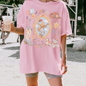 Puede incluir: Una camiseta rosa claro con un estampado gr&aacute;fico central de una persona tocando la guitarra dentro de un marco dorado ornamentado, rodeada de ilustraciones de una bota vaquera, un sombrero, una estrella de sheriff, un oso de peluche, un caballito de juguete y globos.