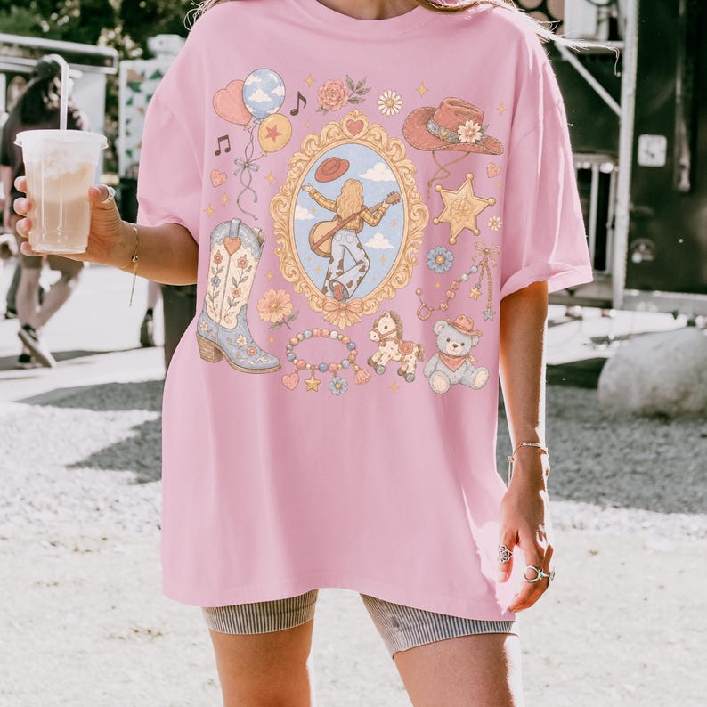 Puede incluir: Una camiseta rosa claro con un estampado gr&aacute;fico central de una persona tocando la guitarra dentro de un marco dorado ornamentado, rodeada de ilustraciones de una bota vaquera, un sombrero, una estrella de sheriff, un oso de peluche, un caballito de juguete y globos.
