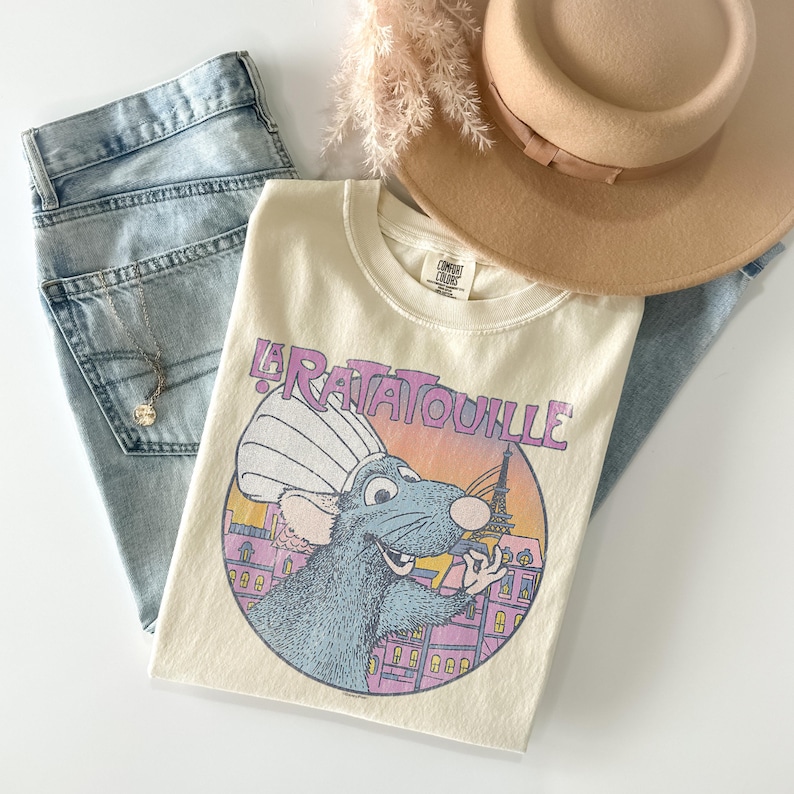 May include: Cream-colored t-shirt featuring a cartoon illustration of a rat chef from the movie "Ratatouille" with the Eiffel Tower in the background. The shirt is paired with light-wash denim jeans, a tan hat, and a necklace.
