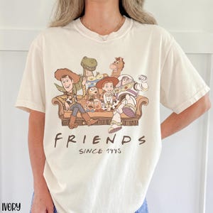 May include: Ivory t-shirt with a graphic of Toy Story characters on a brown sofa. The word "FRIENDS" is printed below the graphic, with "SINCE 1995" underneath. The shirt has short sleeves and a crew neck.