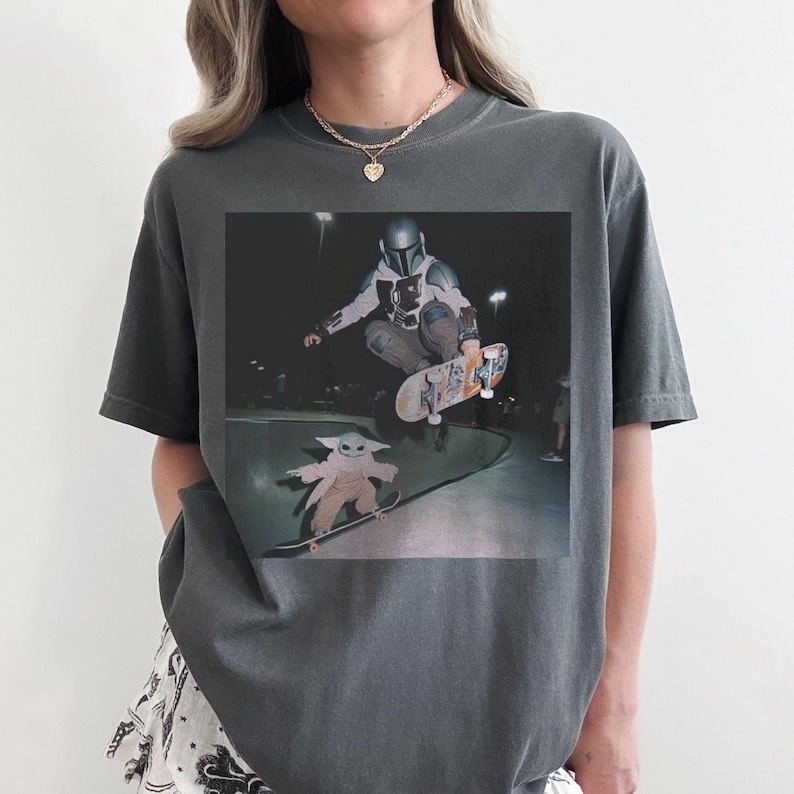 Pu&ograve; includere: Una t-shirt girocollo grigio antracite con una stampa grafica che ritrae un personaggio mandaloriano mentre esegue un trick con lo skateboard in aria sopra una piccola creatura verde su uno skateboard in uno skatepark in cemento di notte.