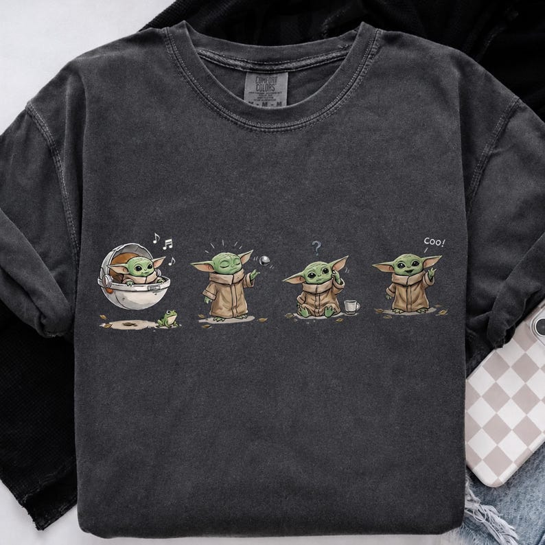 May include: A charcoal gray crew neck t-shirt featuring a graphic print of a small green creature in four different poses. The illustrations show the character in a floating pod, standing, sitting with a question mark, and waving with the text coo.