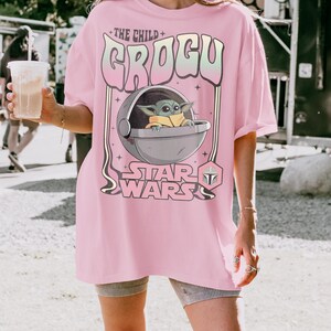 K&ouml;nnte beinhalten: Ein hellrosa T-Shirt mit einem Grafikdruck von Grogu aus Star Wars in einer schwebenden Kapsel. Der Text lautet THE CHILD GROGU und STAR WARS, erg&auml;nzt durch ein kleines Mandalorianer-Helm-Symbol. Das Design verwendet eine retro, wellenf&ouml;rmige Schrift.