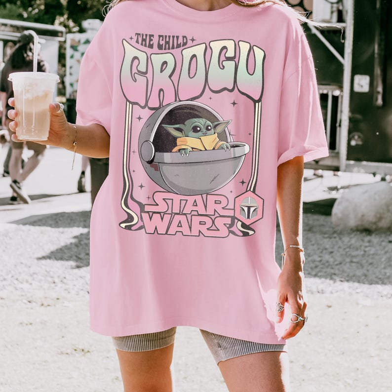 K&ouml;nnte beinhalten: Ein hellrosa T-Shirt mit einem Grafikdruck von Grogu aus Star Wars in einer schwebenden Kapsel. Der Text lautet THE CHILD GROGU und STAR WARS, erg&auml;nzt durch ein kleines Mandalorianer-Helm-Symbol. Das Design verwendet eine retro, wellenf&ouml;rmige Schrift.