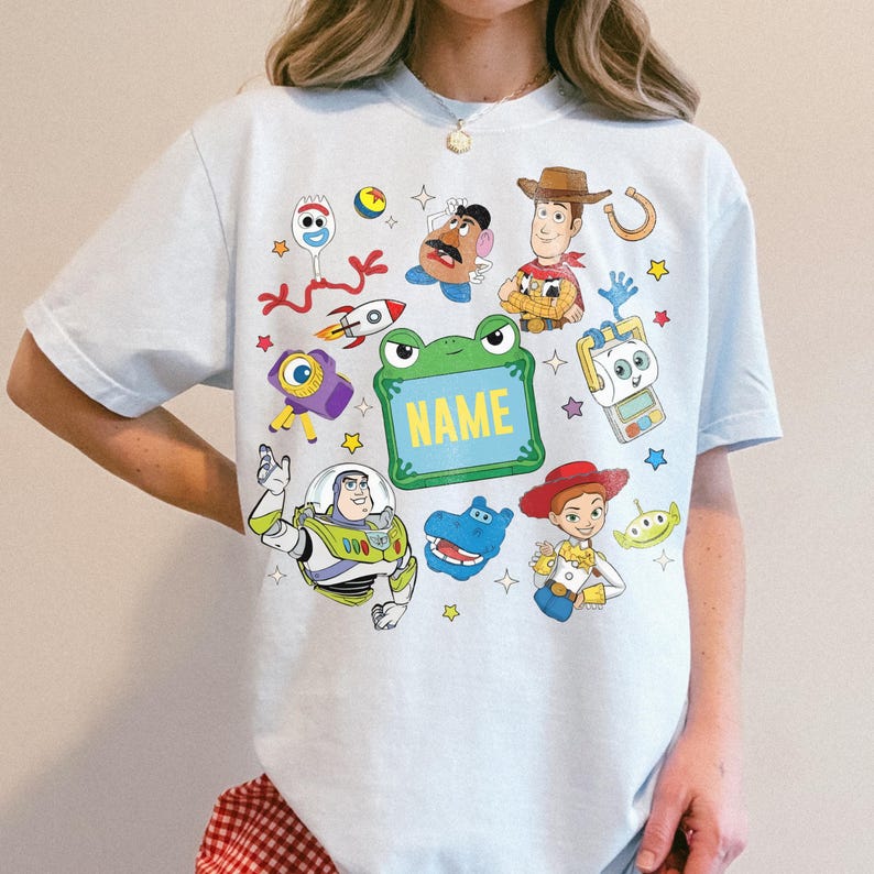 May include: A light blue t-shirt featuring a graphic print of various animated characters including a cowboy, a space ranger, a cowgirl, a potato figure, and a fork character, surrounding a central green frame with the word NAME printed in yellow.