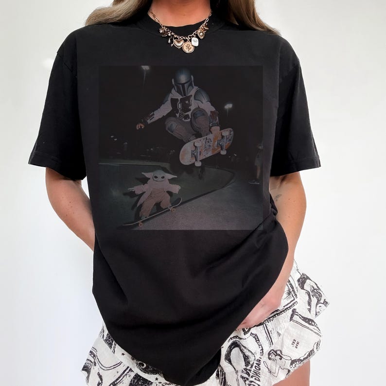 Pu&ograve; includere: Una t-shirt nera a maniche corte con una stampa grafica che ritrae un personaggio in armatura argentata mentre esegue un trick con lo skateboard in aria, mentre una piccola creatura verde sta su un altro skateboard a terra.