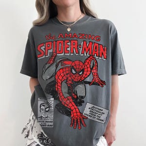 May include: A charcoal gray short-sleeve t-shirt featuring a distressed red and black graphic of Spider-Man. The text reads "the AMAZING SPIDER-MAN" at the top, with a small "PETER PARKER ALIAS SPIDER-MAN" box and a "ANOTHER SPECTACULAR SMASH-HIT" text box.