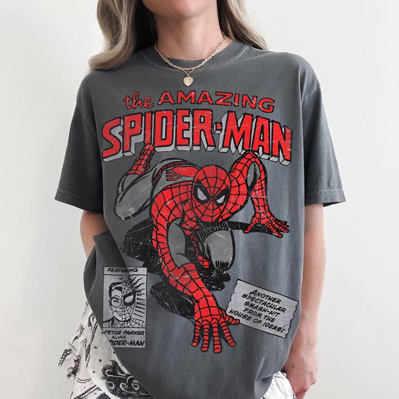May include: A charcoal gray short-sleeve t-shirt featuring a distressed red and black graphic of Spider-Man. The text reads "the AMAZING SPIDER-MAN" at the top, with a small "PETER PARKER ALIAS SPIDER-MAN" box and a "ANOTHER SPECTACULAR SMASH-HIT" text box.