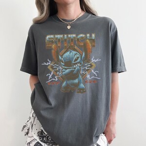 May include: Dark gray t-shirt featuring the character Stitch from Lilo & Stitch. The design includes the word "STITCH" in a retro font, Stitch himself, and the text "626". The graphic is surrounded by lightning bolts.