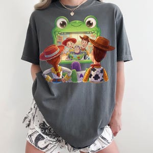 Puede incluir: Camiseta gris oscuro con un gr&aacute;fico colorido de personajes de la pel&iacute;cula animada "Toy Story". El dise&ntilde;o muestra a Woody, Buzz Lightyear, Jessie y otros personajes mirando una pantalla. La camiseta es de corte holgado.