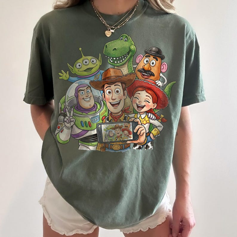 May include: Olive green t-shirt featuring a graphic of Toy Story characters taking a selfie. The design includes Buzz Lightyear, Woody, Jessie, Rex, Mr. Potato Head, and an alien. The characters are smiling and holding a phone.