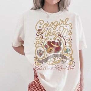May include: An off-white short-sleeve t-shirt featuring a graphic print with the text Cowgirl Like Me and Takes One to Know One. The design includes a cowboy hat, a horseshoe, a sun, and rope motifs in shades of red, yellow, and brown.