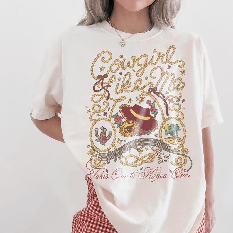 May include: An off-white short-sleeve t-shirt featuring a graphic print with the text Cowgirl Like Me and Takes One to Know One. The design includes a cowboy hat, a horseshoe, a sun, and rope motifs in shades of red, yellow, and brown.