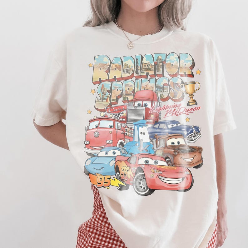 Op de afbeelding: Een gebroken wit t-shirt met korte mouwen en een grafische print van personages uit de film Cars. De tekst Radiator Springs en Lightning McQueen staat boven een verzameling kleurrijke geanimeerde voertuigen, waaronder een rode raceauto.