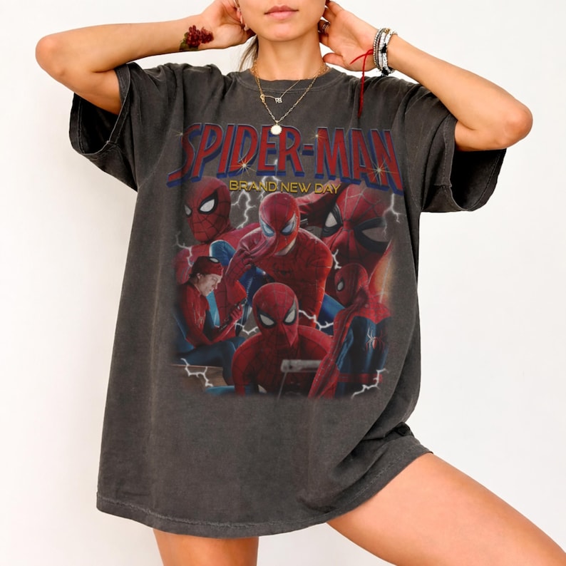May include: A charcoal gray oversized t-shirt featuring a graphic print of Spider-Man in various poses. The text "SPIDER-MAN" appears in bold red letters at the top, with "BRAND NEW DAY" printed underneath. The design includes lightning bolt accents.