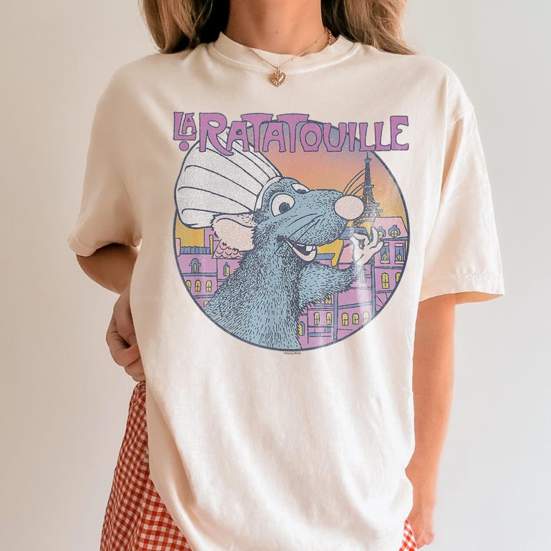 May include: Cream-colored t-shirt featuring a circular graphic from the movie "Ratatouille." The graphic shows a blue rat wearing a chef's hat, with the Eiffel Tower in the background and the words "La Ratatouille" above.