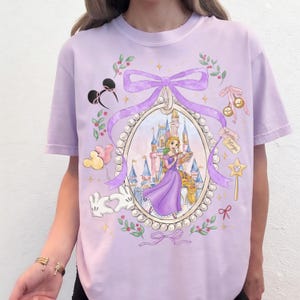 Pu&ograve; includere: Una maglietta lilla con una principessa che dipinge un castello. Il design include una cornice ovale bordata di perle, un fiocco, orecchie di Topolino, palloncini e una bacchetta magica. Le parole "Magic Magic" sono su un'etichetta.