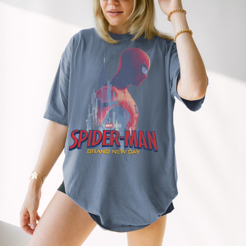 May include: A slate blue oversized t-shirt featuring a graphic of Spider-Man in a red suit against a city skyline. The text reads Marvel Studios Spider-Man Brand New Day in red and yellow lettering. The shirt has short sleeves and a crew neckline.