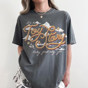 May include: A charcoal gray short-sleeve t-shirt featuring a graphic print of white clouds and the text "it's a Toy Story" written in a rope-style font. Below the main text, the phrase "Baby just say yes" is printed in a white cursive script.