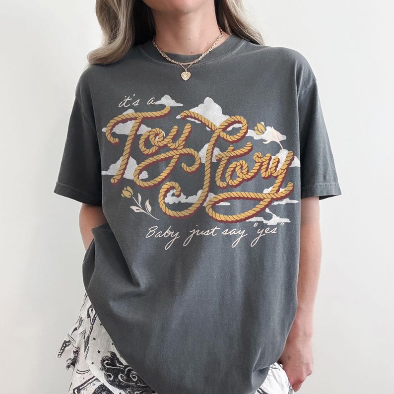 May include: A charcoal gray short-sleeve t-shirt featuring a graphic print of white clouds and the text "it's a Toy Story" written in a rope-style font. Below the main text, the phrase "Baby just say yes" is printed in a white cursive script.