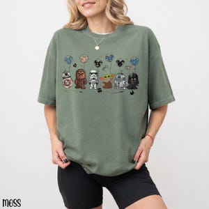 May include: Sage green t-shirt featuring a cartoon graphic of Star Wars characters as balloons. The characters include BB-8, Chewbacca, a Stormtrooper, Grogu, R2-D2, and Darth Vader. The word "moss" is printed in the bottom left corner.