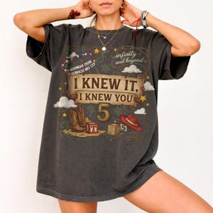 May include: A charcoal gray short-sleeve t-shirt featuring a graphic print with the text "I KNEW IT, I KNEW YOU", "infinity and beyond", and "YOU'VE GOT A FRIEND IN ME". The design includes a cowboy hat, boots, blocks, and stars.