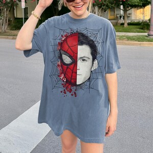 May include: A slate blue short-sleeve t-shirt featuring a graphic print of a split face. One side shows a red and black spider-themed mask with web patterns and red paint splatters, while the other side displays a black and white portrait of a human.