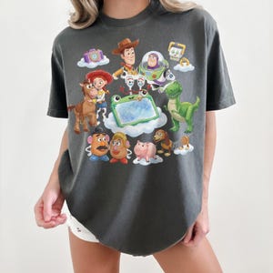 May include: Dark gray t-shirt featuring colorful illustrations of characters from the animated movie Toy Story. The design includes Woody, Buzz Lightyear, and other characters on a cloud background. The shirt has a crew neck and short sleeves.