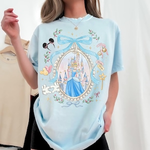 Pu&ograve; includere: T-shirt azzurra con un motivo a tema Cenerentola. Il design include un ritratto di Cenerentola in un abito blu, un castello e altri elementi Disney. Ulteriori dettagli includono un fiocco, una bacchetta magica e le parole "Minnie Magic".
