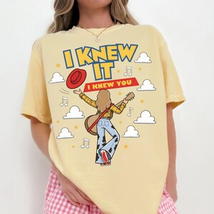 May include: A pale yellow t-shirt featuring a graphic illustration of a person with long hair playing an acoustic guitar, wearing a yellow plaid shirt and blue cow-print bell-bottom pants. The text reads I KNEW IT and I KNEW YOU, with clouds, stars, and musical notes.